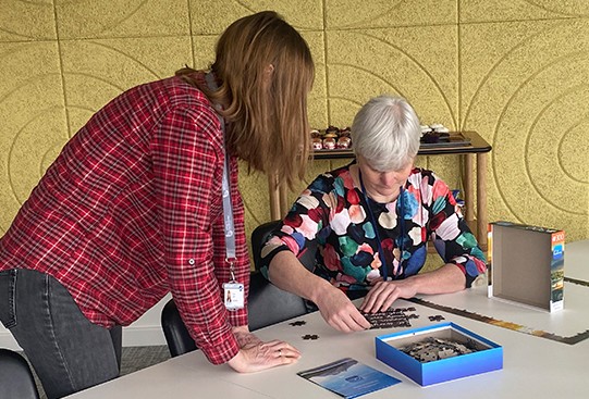 Two women are engaged in assembling a jigsaw puzzle on a table, one sorting pieces while the other focuses on placing them, surrounded by a bright, modern room with a textured yellow wall.