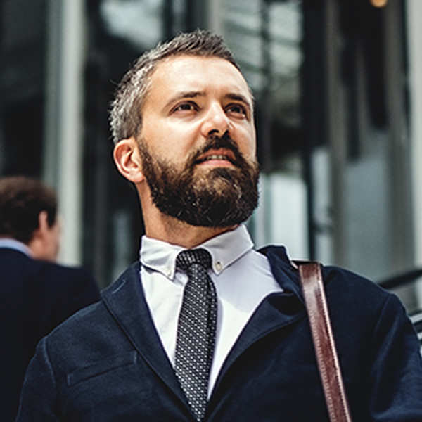 A bearded man in a suit confidently ascends an escalator, looking forward. A blurred figure in business attire walks past him, suggesting a busy, modern urban environment.