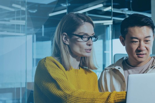 A woman in a yellow sweater and a man in a light jacket collaborate over a laptop in a modern office setting, surrounded by glass walls and plants, actively discussing something on the screen.