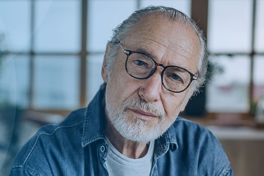 An older man with glasses sits thoughtfully in a well-lit room, surrounded by plants and a bicycle, conveying a sense of contemplation and wisdom.