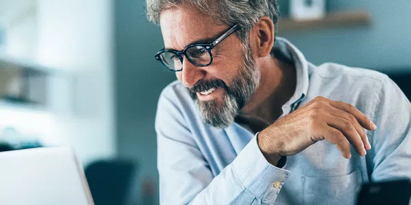 A man with glasses smiles while using a laptop, engaged and focused. The setting is a modern workspace with soft lighting, suggesting a relaxed atmosphere conducive to concentration.