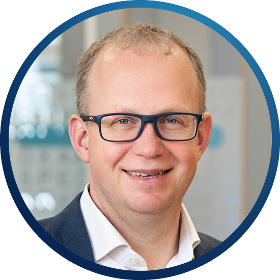 A smiling man with glasses and short hair is positioned in a circular frame. He is wearing a dark suit with a white shirt, set against a blurred office background.