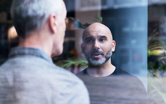 A man with a bald head and an expressive face is conversing while standing behind a glass door, reflecting a blurred figure of another person, set in a modern interior surrounded by plants.