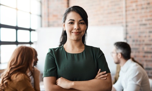 A woman with long dark hair stands confidently with her arms crossed, smiling, in a modern workspace with colleagues blurred in the background, featuring exposed brick walls and large windows.