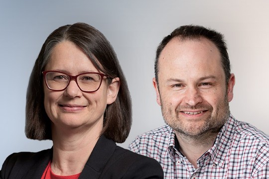 Two professionals are smiling, one with glasses and straight hair, the other with a beard and a checkered shirt. They are posed against a light, neutral background.