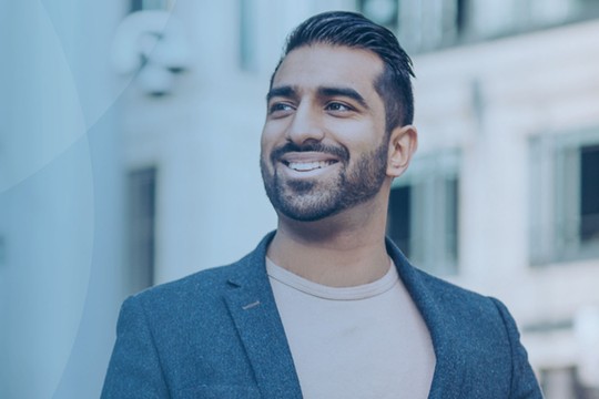A man stands confidently, smiling while wearing a blazer over a light-colored shirt. The background features blurred contemporary buildings, suggesting an urban environment.