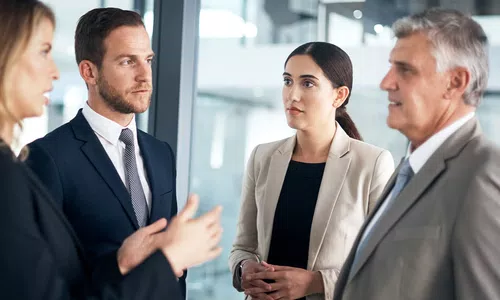 Four professionals engage in a discussion. The setting is an office space with glass elements. They wear formal attire, indicating a business context, and display attentive expressions.