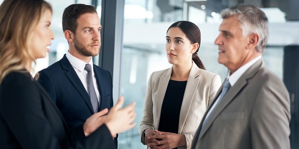 Four professionals engage in a discussion. The setting is an office space with glass elements. They wear formal attire, indicating a business context, and display attentive expressions.