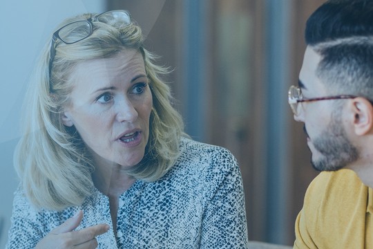 A woman with blonde hair and glasses gestures while discussing with a man in a yellow shirt, both seated at a table in a softly lit, modern environment.