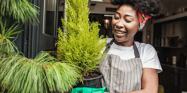 A woman with curly hair, wearing a striped apron and green gloves, smiles while holding a potted green plant, standing near a door with a warm interior background.