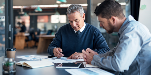 Two men are seated at a table discussing documents covered with graphs and charts in a modern office setting, with a coffee cup and a notebook nearby.