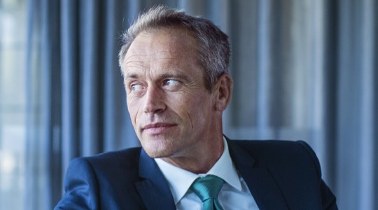 A man in a dark suit and teal tie sits pensively by a large window, gazing thoughtfully outside. Soft gray curtains and glass elements enhance the modern office environment.