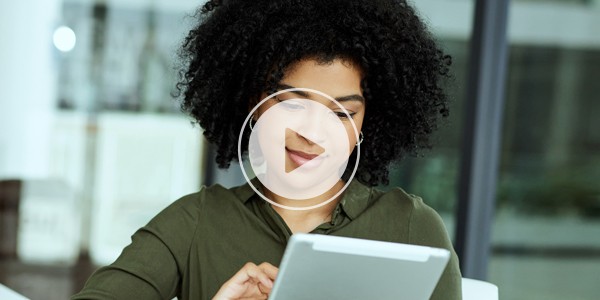 A woman with curly hair is focused on a tablet, engaging with its screen. She is seated indoors, surrounded by a bright, modern environment featuring large windows.