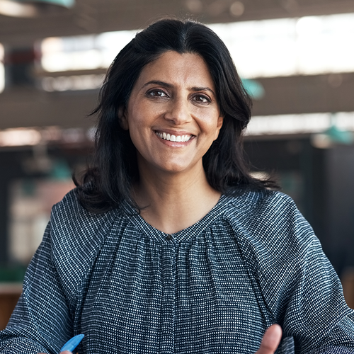 A woman with long dark hair smiles warmly while seated at a table, holding a pen. The background features soft lighting and hints of a workspace environment.