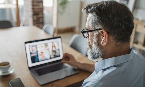 A man with a beard uses a laptop, engaging in a video call while wearing wireless earbuds. He sits at a wooden table with a cup of coffee and a smartphone nearby, in a bright, casual setting.