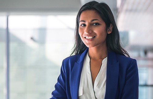 A smiling woman in a blue blazer stands, engaging with the viewer. She has long, dark hair and is positioned in a well-lit, modern interior with blurred background elements.
