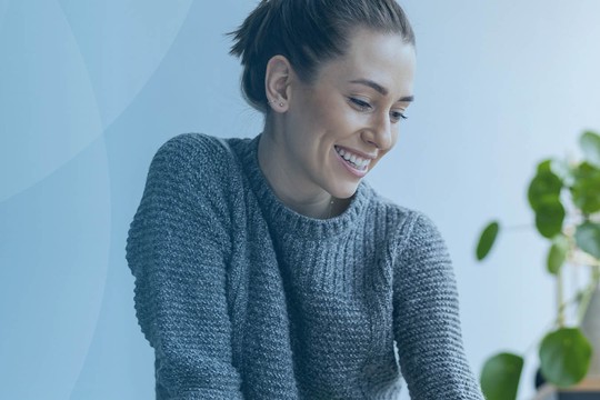 A woman smiles while engaging with an object on a table, surrounded by a softly lit, modern indoor space with plants and large windows, creating a calm atmosphere.
