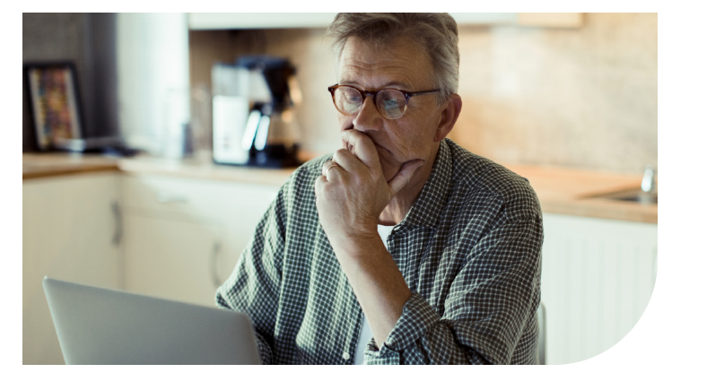 A man sits at a table, focused on his laptop while resting his chin on his hand. A coffee maker is visible in a well-lit kitchen behind him.