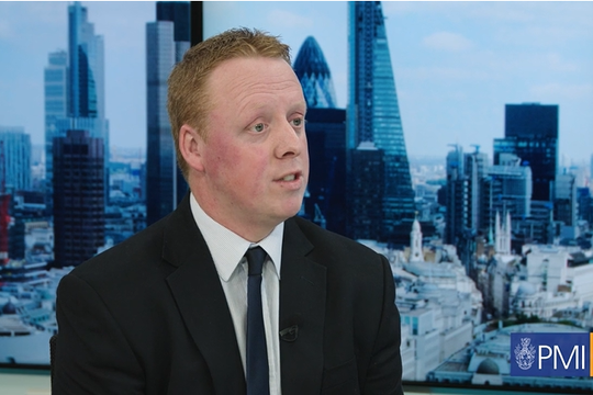 A man in a suit is speaking, likely during an interview. Behind him is a city skyline featuring modern buildings and a clear sky, suggesting a professional or business setting.