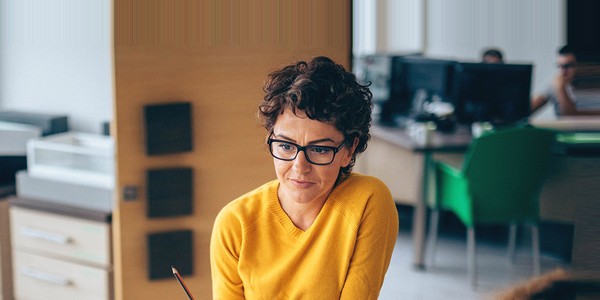 A woman wearing a yellow sweater and glasses sits thoughtfully, holding a pencil. She is in an office space with desks and computers in the background.