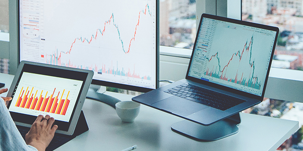 A person is analyzing data on a tablet and laptop, surrounded by financial charts on screens, with a cityscape visible through large windows in a modern office setting.