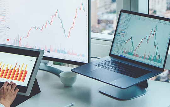 A person is analyzing data on a tablet and laptop, surrounded by financial charts on screens, with a cityscape visible through large windows in a modern office setting.