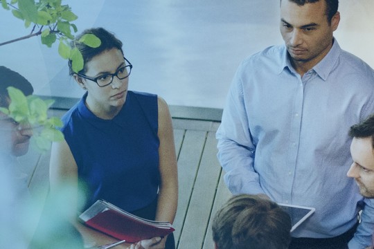 A group of five professionals are engaged in discussion, holding documents and looking at each other. They are surrounded by natural light and greenery in a modern, indoor setting.