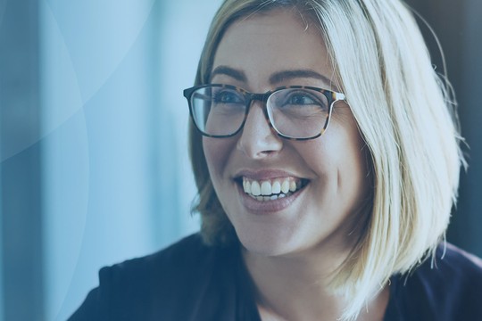 A smiling woman with short blonde hair and glasses is sitting indoors, illuminated by natural light coming through a window, against a soft blue background.