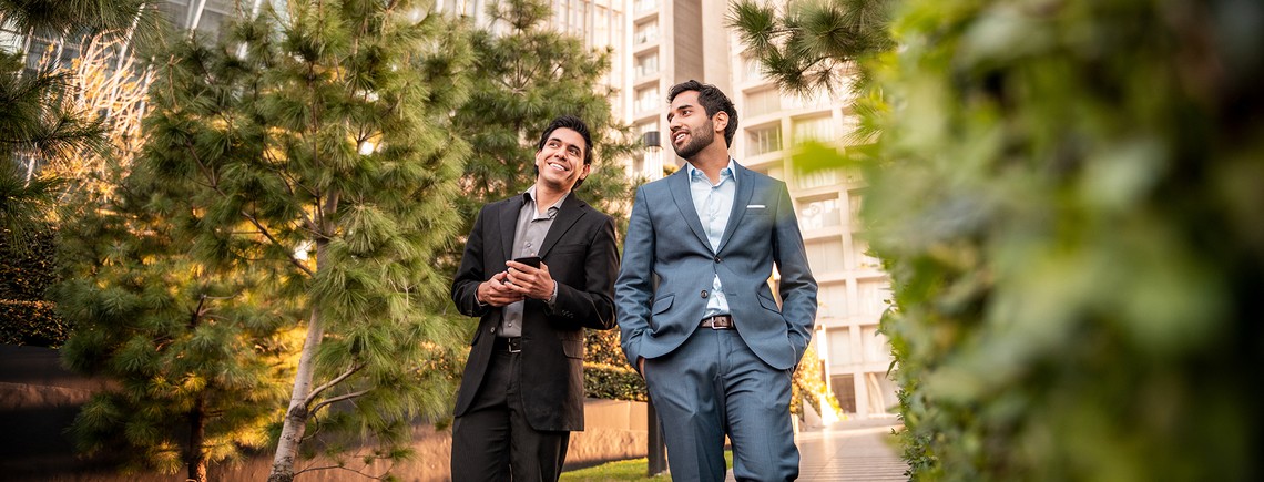 Two men in professional attire walk together through a landscaped area with trees and modern buildings. One man is looking at his phone, both appear to be enjoying the moment.