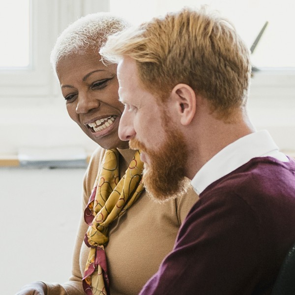 A smiling woman and a man engaged in conversation share ideas while sitting close together in a well-lit room, surrounded by minimal furnishings and a bright window.