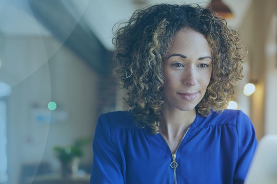 A woman with curly hair in a blue top is focused on her laptop in a cozy, well-lit indoor space featuring plants and warm lighting, suggesting a relaxed, productive setting.
