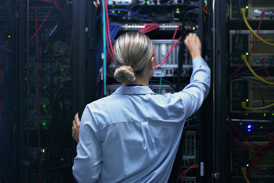 A person in a light blue shirt adjusts equipment inside a server rack. Colorful cables connect various devices, with blinking indicator lights in a dimly lit data center.