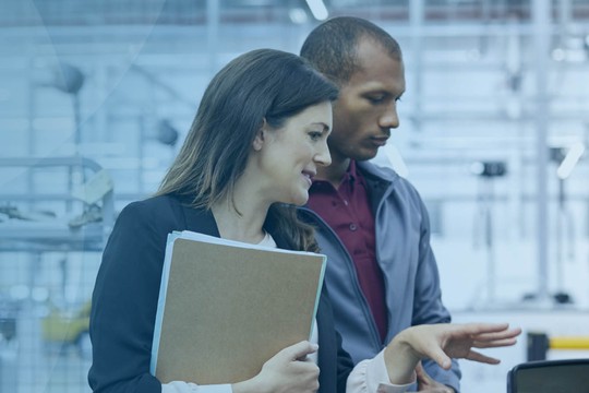 A man and a woman are discussing a car in a well-lit industrial or workshop environment. The woman holds a clipboard, gesturing, while the man listens attentively.