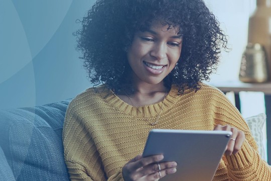 A woman with curly hair sits on a couch, smiling while using a tablet. She is dressed in a yellow sweater, surrounded by a cozy living room with decorative items in the background.