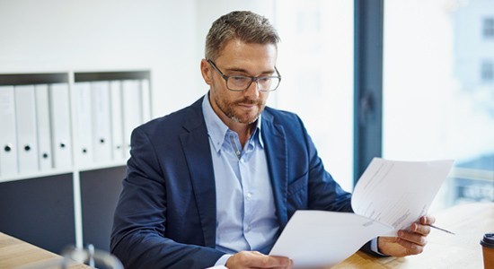 A man in a business suit reads documents at a desk, holding a pen, with shelves of files and a large window in a modern office setting.