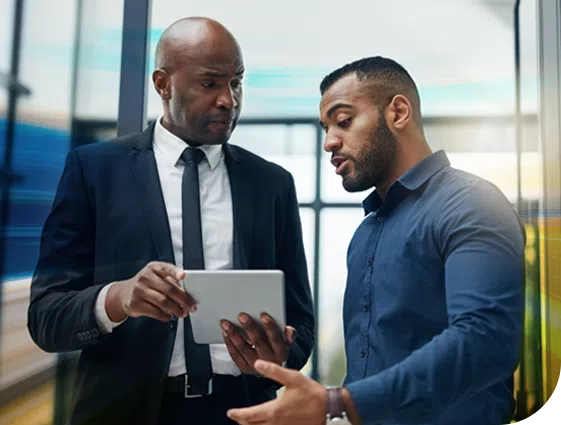 Two men discuss information displayed on a tablet. They stand in a modern office with large windows, suggesting a professional environment. One man wears a suit, the other a casual shirt.