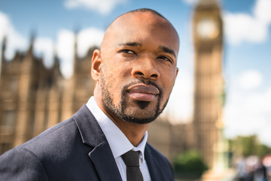 A man in a suit stands confidently, gazing into the distance. The backdrop features the iconic Big Ben clock tower and historical buildings under a clear blue sky with scattered clouds.