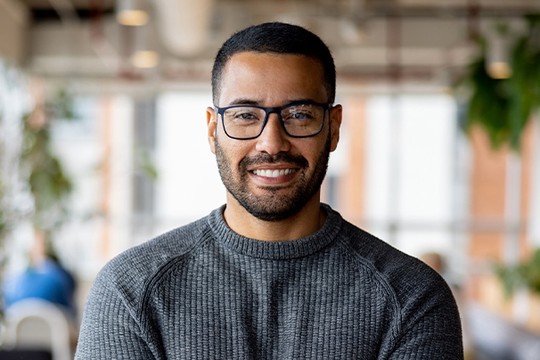 A man with glasses and short hair smiles confidently while standing in a modern office space, featuring large windows and greenery, suggesting a collaborative environment.