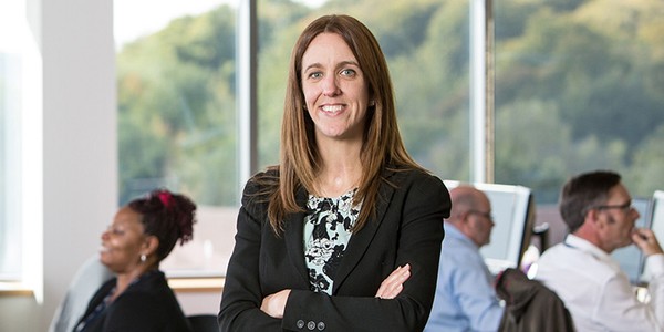 A woman in a black blazer stands confidently with arms crossed, smiling. In the background, several people work at desks, with large windows revealing a lush green landscape outside.