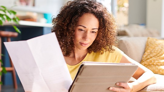 A woman sits on a sofa, reading a document while using a tablet. She is in a bright, cozy living room with plants and soft furnishings, suggesting a relaxed atmosphere.