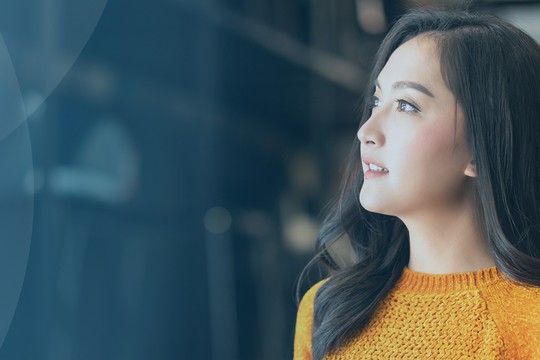 A woman in an orange sweater stands, looking thoughtfully to the side. Soft blue tones and a blurred background suggest a modern, indoor environment, creating a calm atmosphere.