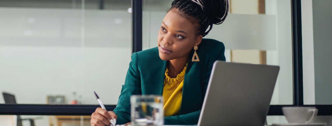 A woman in a green blazer focuses intently while writing with a pen. A laptop and a glass of water are on the table in a modern office setting.