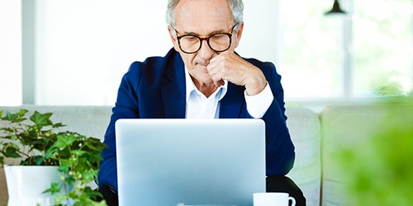 A middle-aged man in a blue blazer sits at a table, focused on his laptop. Surrounding him are potted plants and stacks of books, with a coffee cup nearby in a bright, airy room.