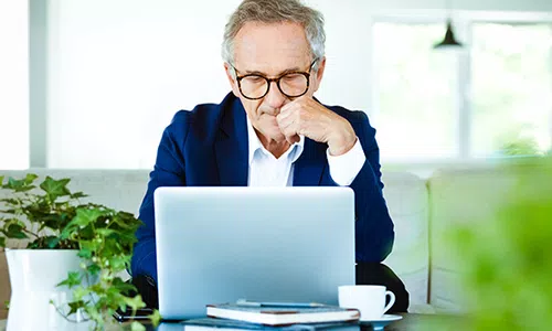 A middle-aged man in a blue blazer sits at a table, focused on his laptop. Surrounding him are potted plants and stacks of books, with a coffee cup nearby in a bright, airy room.