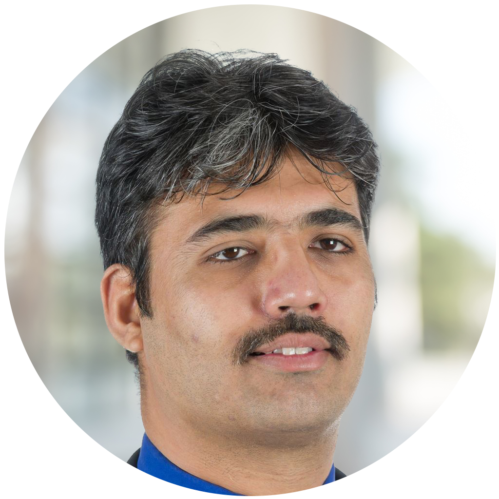 A man with dark, slightly wavy hair and a mustache is looking directly at the camera, dressed in a blue shirt and dark blazer, set against a softly blurred background.