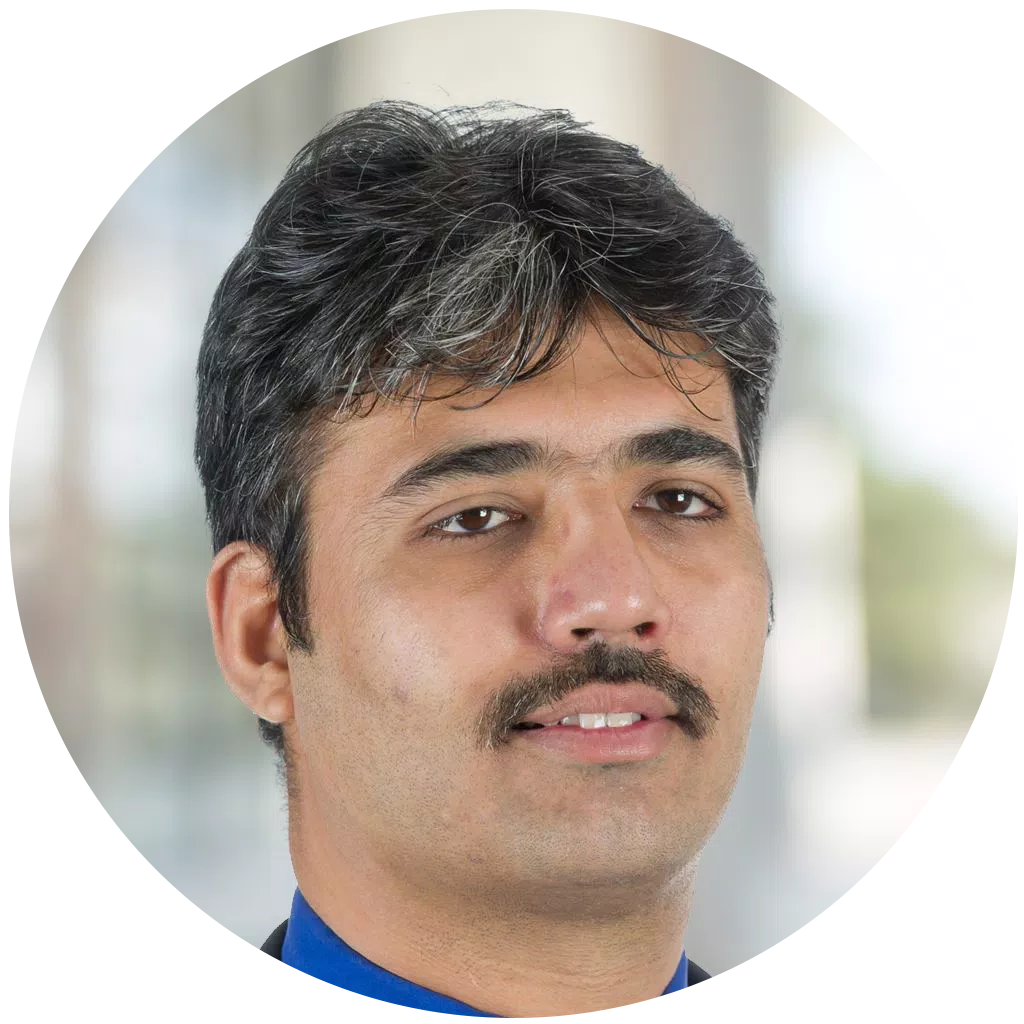A man with dark, slightly wavy hair and a mustache is looking directly at the camera, dressed in a blue shirt and dark blazer, set against a softly blurred background.