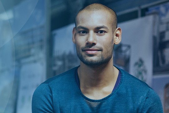 A man wearing a blue shirt sits calmly, facing the camera. Behind him are blurred images of architectural designs, suggesting a creative workspace or studio environment.