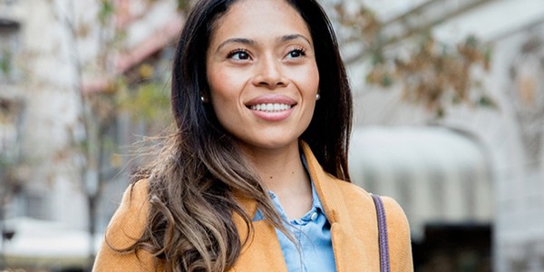 A woman with long, wavy hair smiles while walking outdoors. She wears a yellow coat over a blue shirt in an urban setting with blurred buildings and greenery in the background.