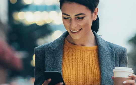 A woman in a gray blazer and yellow sweater smiles while holding a smartphone in one hand and a coffee cup in the other, walking along a city street in soft evening light.