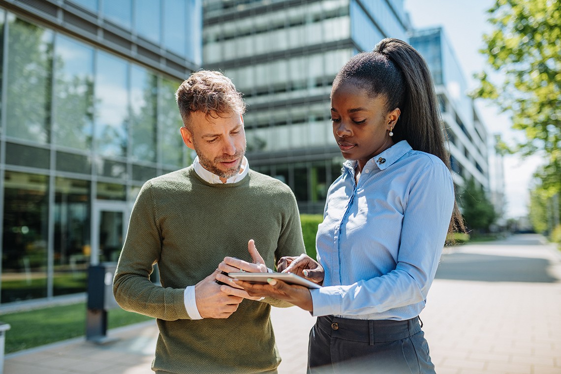 Two professionals, a man and a woman, are engaged in a discussion while examining a tablet. They are outdoors near modern office buildings, under clear skies.